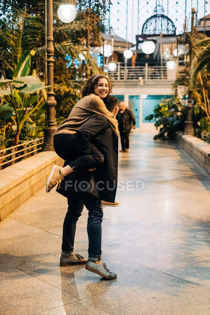 Excited young male giving piggyback ride to cheerful young female while walking along path during romantic date on city street — Stock Photo