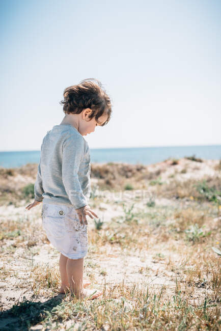 Vista laterale del bambino carino in piedi con i piedi nudi nella sabbia in una bella spiaggia nella giornata di sole — Foto stock