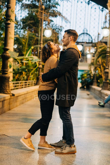 Alegre joven hombre y mujer abrazándose y mirándose mientras están de pie dentro del pabellón iluminado durante la fecha - foto de stock