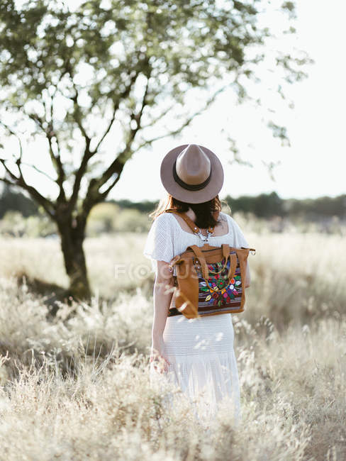 Mulher de chapéu com mochila na moda andando entre campo selvagem à luz do dia — Fotografia de Stock