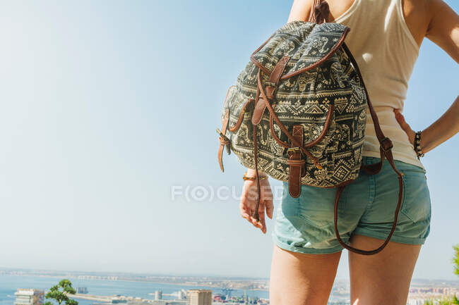Rückenansicht einer jungen Frau in Jeanshosen, die mit ihrem Rucksack über der atemberaubenden Stadtlandschaft und dem ruhigen Wasser bei hellem Tag in Alicante Spanien steht — Stockfoto