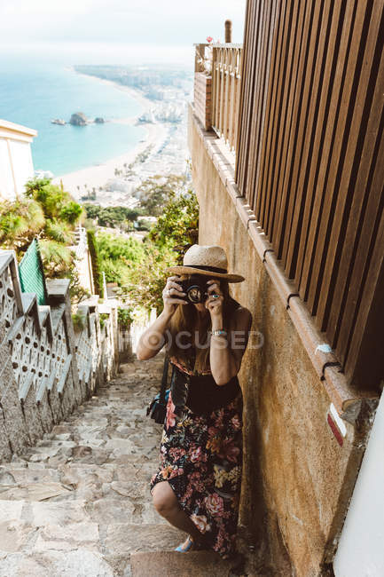 Жінка в літньому вбранні фотографується, стоячи на вулиці кам'яних сходах з морським узбережжям на фоні — стокове фото