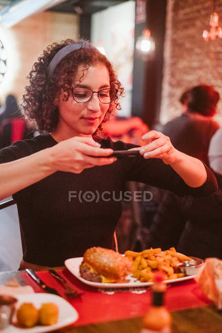 Молода жінка з кучерявим волоссям, використовуючи смартфон, щоб сфотографувати бургер і картоплю фрі, сидячи за столом кафе — стокове фото