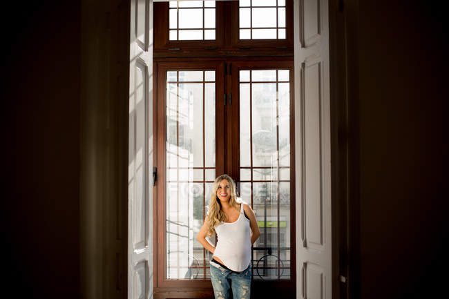Улыбающаяся беременная женщина в повседневной одежде, стоящая у окна дома — стоковое фото
