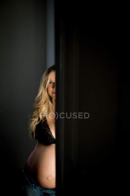 Pregnant woman in bra looking at camera while standing near open door at dark room — Stock Photo