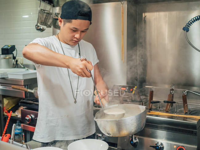 Junger Mann mit schwarzer Mütze gießt heiße Brühe aus Topf für japanisches Gericht namens Ramen in asiatischem Café — Stockfoto