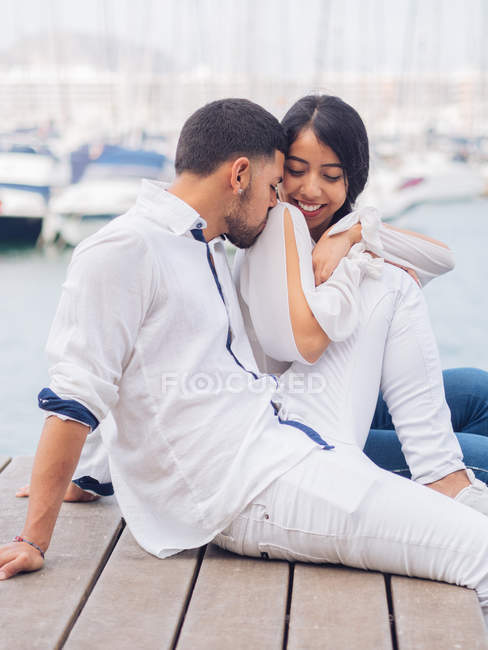 Молодой красавец обнимает и целует привлекательную улыбающуюся женщину, сидящую на деревянной поверхности — стоковое фото
