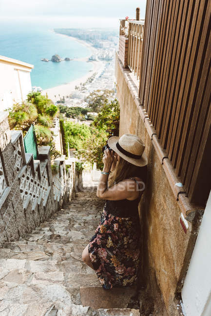 Frau im Sommer-Outfit beim Fotografieren, während sie auf einer Steintreppe mit Meeresküste im Hintergrund steht — Stockfoto