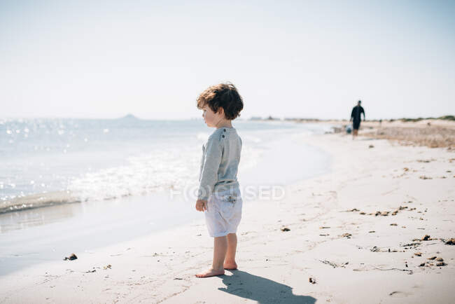 Vista lateral de bebé lindo de pie con los pies desnudos en la arena y mirando el mar tranquilo brillante en la hermosa playa en el día soleado - foto de stock
