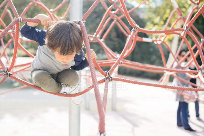 Niño relajado pensativo colgando de la red de escalada de cuerda en el patio de recreo en luz brillante - foto de stock