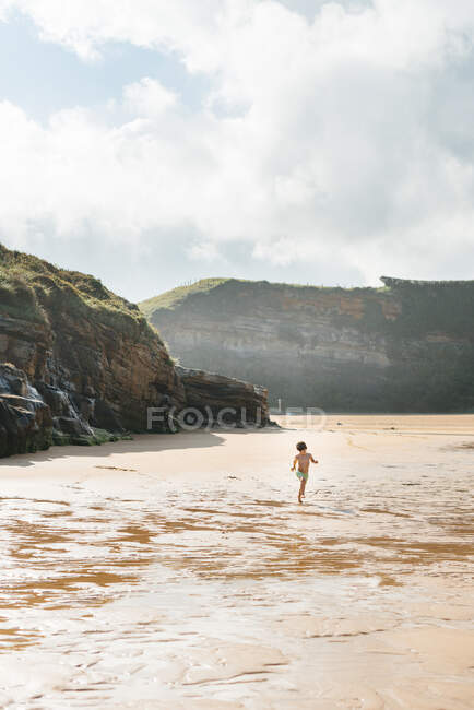 Niño caminando sobre la arena mojada de la orilla del mar a la luz del sol en una playa rocosa - foto de stock
