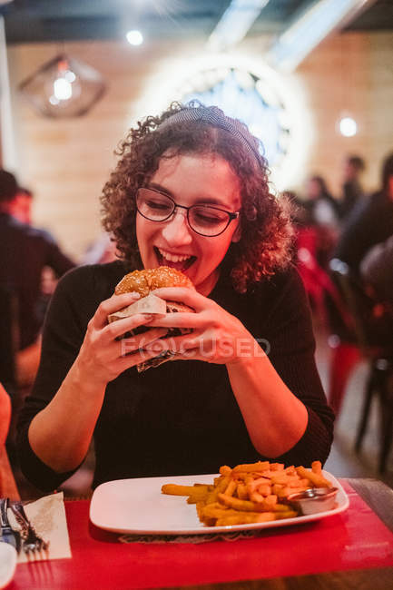 Hungry young woman biting tasty burger while sitting at table in brightly illuminated cafe — Stock Photo