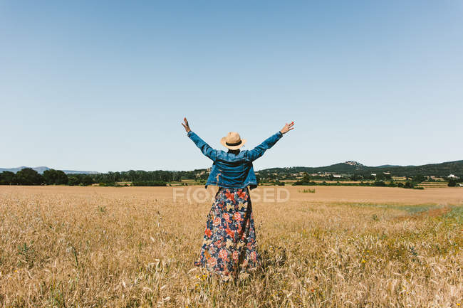 Back view of unrecognizable woman in hat standing on wheat field with outstretching arms on sunny day — Stock Photo