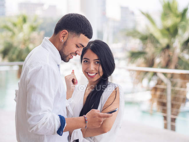 Молода приваблива жінка в білому одязі посміхається дивитися на камеру, коли чоловік пише повідомлення на мобільний телефон — стокове фото