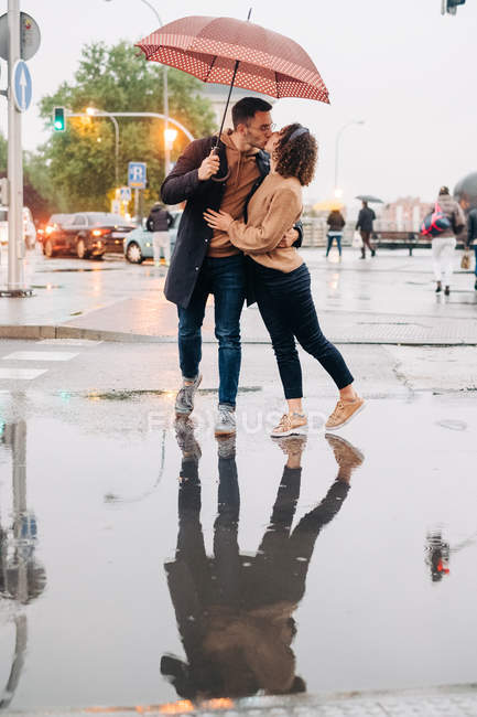Весёлый молодой человек и женщина с зонтиком обнимаются и смотрят друг на друга, стоя на мокрой городской улице в дождливый день — стоковое фото