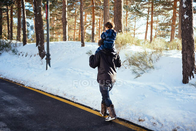 Side view of young man in dark clothes holding child in overall on neck and walking on yellow stripe on road along snowy forest — Stock Photo