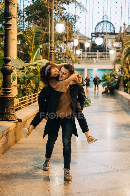 Emocionado jovem do sexo masculino dando passeio de piggyback para alegre jovem do sexo feminino enquanto caminhando ao longo do caminho durante a data romântica na rua da cidade — Fotografia de Stock