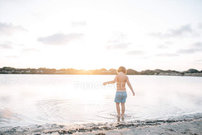 Visão traseira do menino sonhador andando em águas rasas da praia contra a luz do pôr do sol — Fotografia de Stock