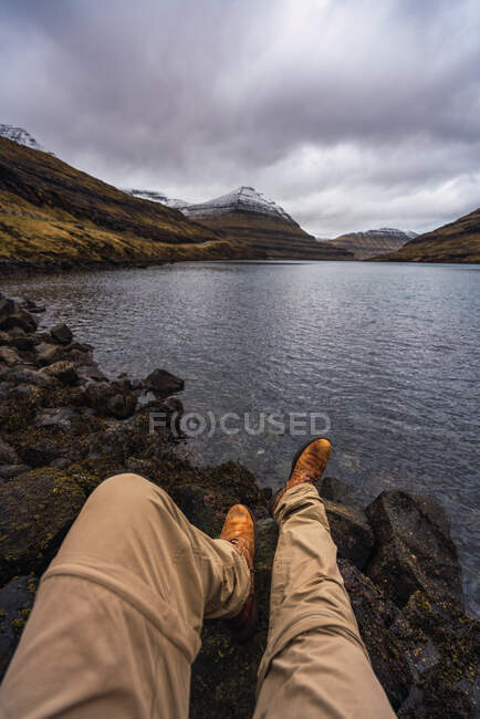 Persona irreconocible sentada cerca del lago mostrando piernas relajándose en la isla de Feroe - foto de stock