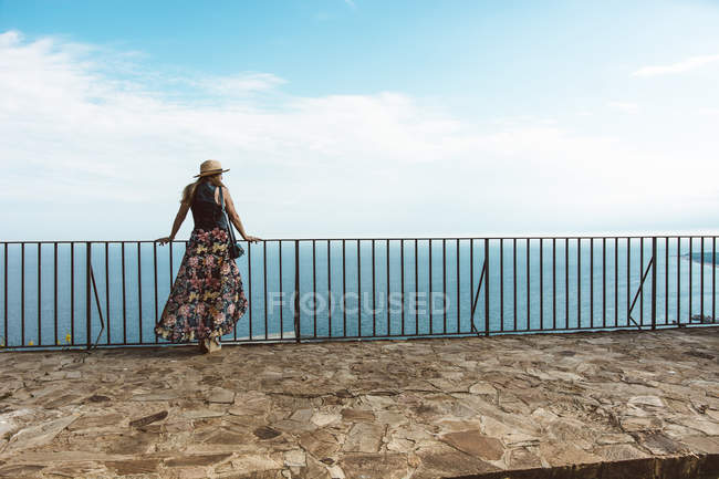 Mujer en vestido de verano y sombrero de pie en la valla en la terraza de piedra con paisaje marino en el fondo - foto de stock