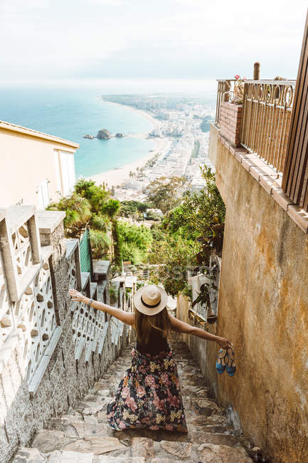 Mujer irreconocible en traje de verano de pie en las escaleras de piedra de la calle con la costa del mar en el fondo - foto de stock