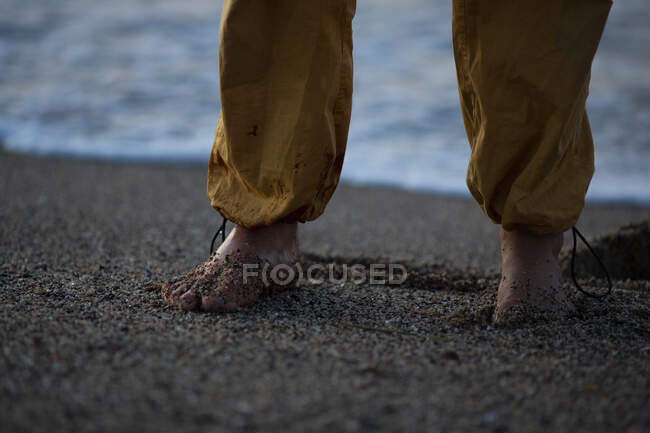 De baixo de pés descalços de pessoa em calças amarelas em pé na areia molhada à beira-mar — Fotografia de Stock