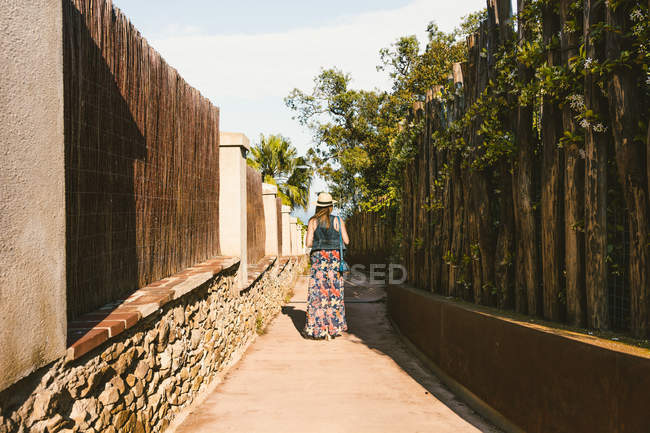 Вид ззаду на невпізнавану жінку в солом'яному капелюсі, що йде на вузькій вулиці на сонці — стокове фото