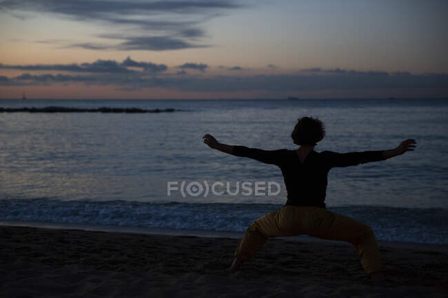 Vista posterior del hombre deportivo en pantalones amarillos descalzo realizando ejercicios de meditación de yoga en la playa de arena en la noche del atardecer - foto de stock