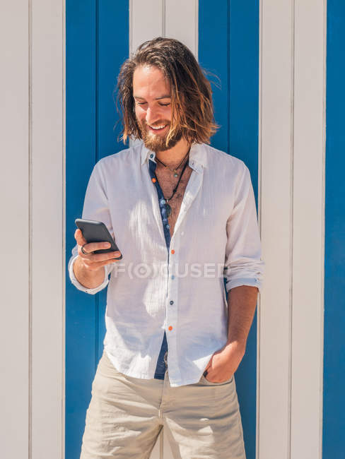 Glücklicher bärtiger Kerl mit Smartphone, während er an einem sonnigen Tag im Resort gegen eine gestreifte Wand steht — Stockfoto