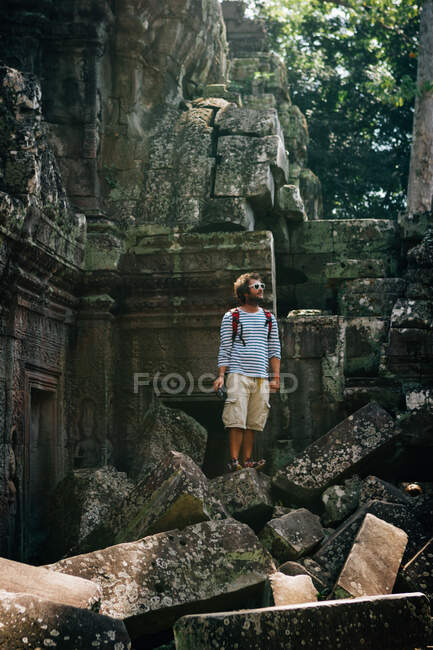 Reisender Mann mit Rucksack, der auf einem Haufen bemooster Steinblöcke steht und antike Tempelruinen erkundet, Thailand — Stockfoto