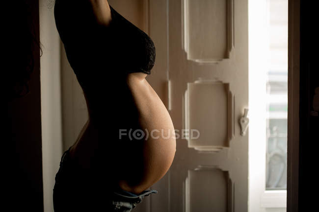 Side view of anonymous pregnant woman in bra standing near open door at home — Stock Photo