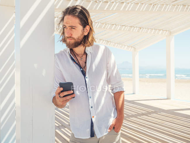 Красивий бородатий чоловік тримає смартфон, спираючись на стовп білої альтанки на піщаному пляжі — стокове фото