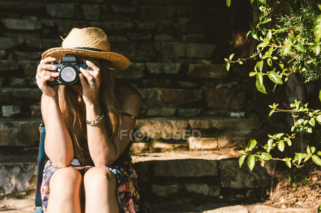 Путешествуя женщина в платье и шляпе с помощью камеры, сидя на каменных ступенях на солнечной улице — стоковое фото