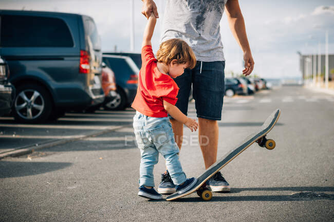 Young man in short holding hands with kid in red t-shirt and helping to skateboard on road in sunny day — Stock Photo