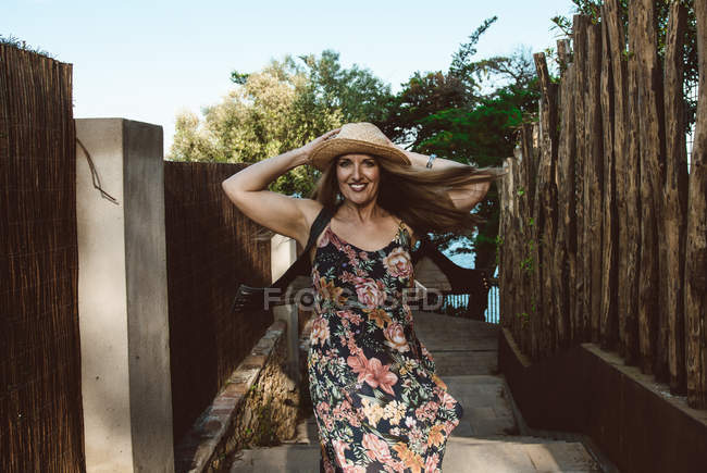 Счастливая взрослая женщина в соломенной шляпе и цветочном платье кружатся на солнечной улице — стоковое фото