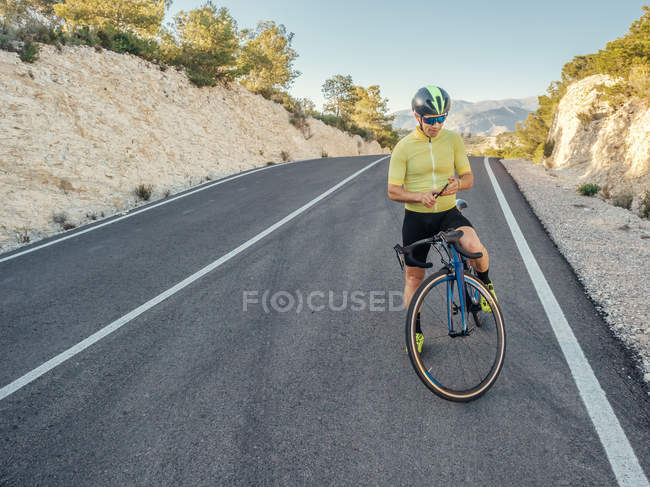 Hombre sano descansando y usando teléfono inteligente con bicicleta en una carretera de montaña en un día soleado - foto de stock