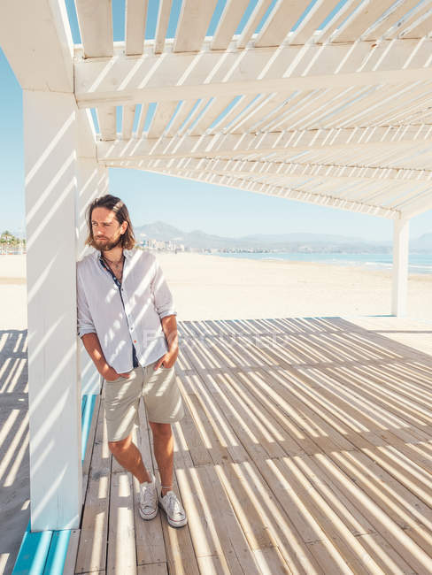 Hombre barbudo guapo mirando hacia otro lado mientras se apoya en el pilar de gazebo blanco en la playa de arena - foto de stock