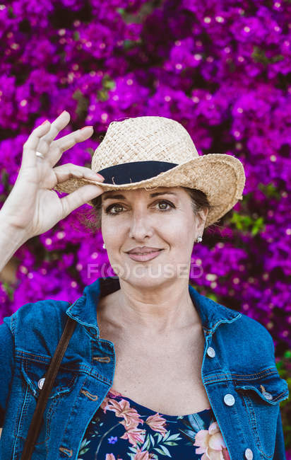 Mulher usando chapéu olhando para a câmera enquanto está perto de flores cor de rosa arbustos — Fotografia de Stock