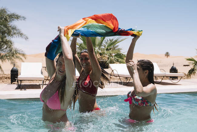 Vista laterale di giovani donne eccitate con bandiera LGBT che si divertono in acqua pulita della piscina — Foto stock