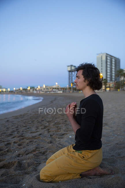 Vista lateral del hombre realizando meditación de yoga en la playa de arena al atardecer - foto de stock