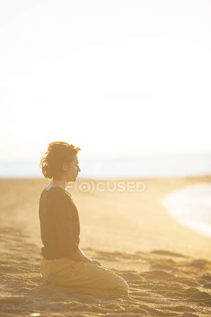 Vista lateral del hombre concentrándose en pensamientos con los ojos cerrados y las manos en gesto de oración sentado de rodillas en la playa de arena a la luz del sol - foto de stock