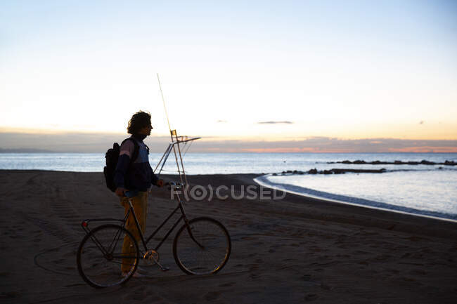 Seitenansicht eines starken jungen Mannes mit Rucksack und Fahrrad am Meer vor dem Hintergrund des Sonnenuntergangs — Stockfoto