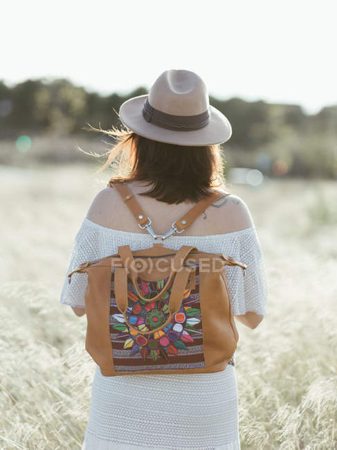 Frau mit Hut und trendigem Rucksack spaziert bei Tageslicht durch wildes Feld — Stockfoto
