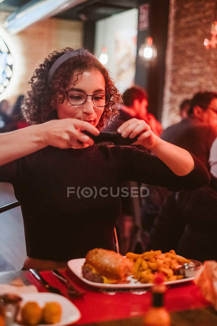 Young woman with curly hair using smartphone to take picture of burger and french fries while sitting at cafe table — Stock Photo