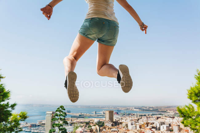 Back view of young woman in denim shorts jumping high above amazing cityscape and calm water in bright day in Alicante Spain — Stock Photo