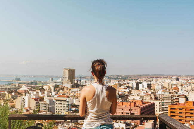 Rückansicht einer jungen Frau im weißen T-Shirt, die bei hellem Tag den Blick vom Balkon auf die Stadt in Alicante genießt — Stockfoto