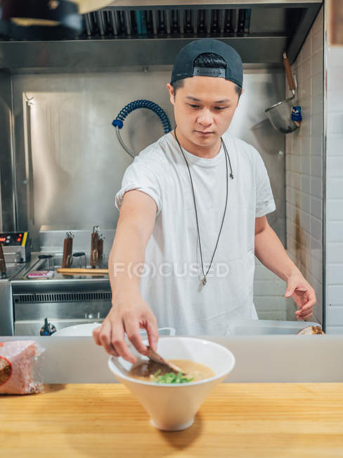 Hombre asiático poniendo huevo en tazón con ramen fresco cocinado en la cocina del restaurante - foto de stock