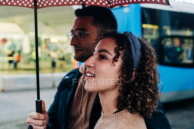Side view of cheerful young man and woman with umbrella walking on wet city street on rainy day — Stock Photo