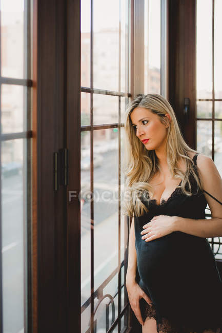Femme enceinte réfléchie en lingerie debout près de la fenêtre à la maison — Photo de stock