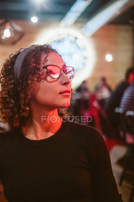 Mujer bastante joven con el pelo rizado y en gafas elegantes mirando hacia otro lado y pensando mientras está sentado en el fondo borroso de la cafetería - foto de stock
